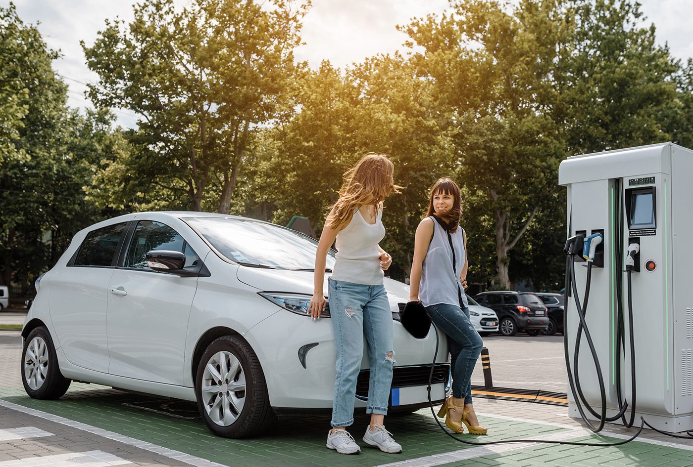 An electric vehicle parked at a charging station, with two women engaged in conversation in front of the car.