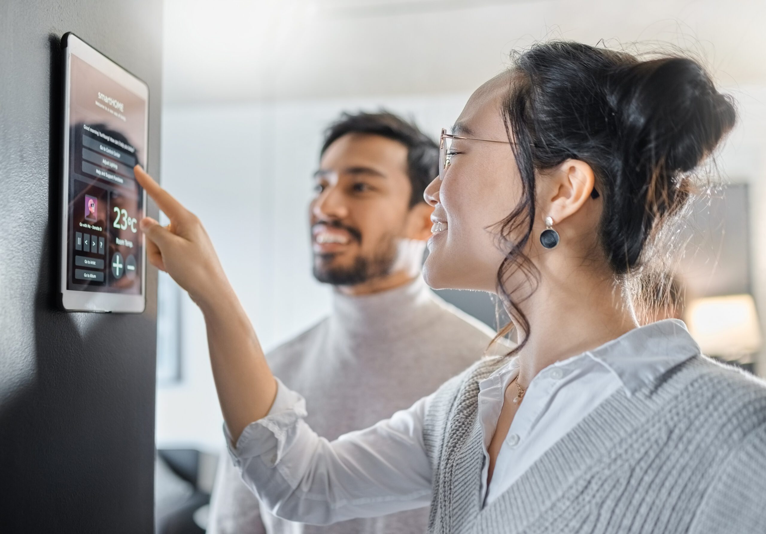 A woman engaging with a smart home touchscreen operating system while her husband stands beside her. The touchscreen displays an intuitive interface for controlling various aspects of the smart home, such as lighting, temperature, and security.